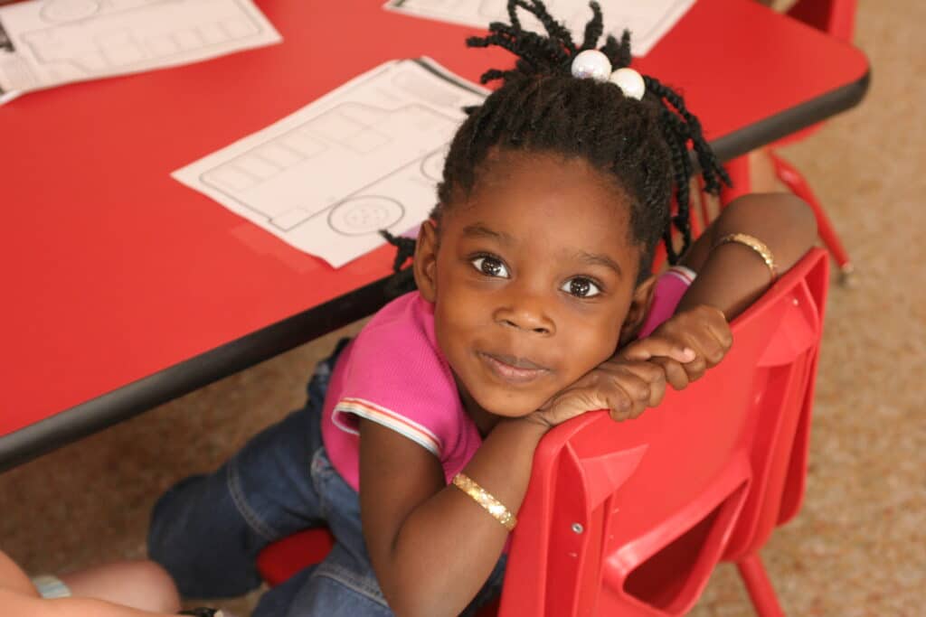 A young child with braided hair and colorful beads rests her chin on her hands, smiling at the camera. She wears a pink shirt and bracelets. She is seated at a red table with paper activities in front of her.