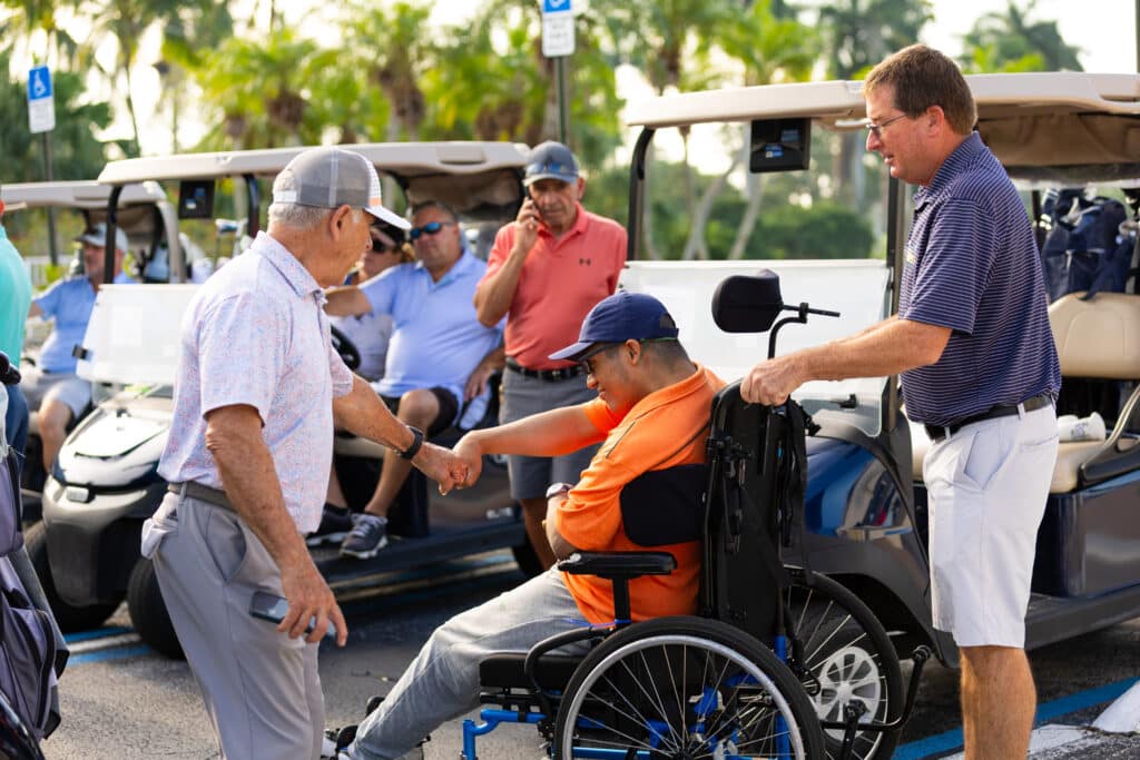 A man in a wheelchair fist-bumps an older man while another man stands beside them. Several people and golf carts are in the background, with palm trees and a sunny sky visible.