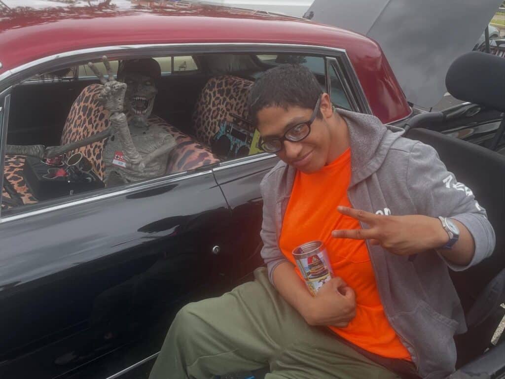 A person in an orange shirt and gray jacket holds a can and flashes a peace sign while posing next to a car with leopard-print seats and a large skeleton decoration inside.