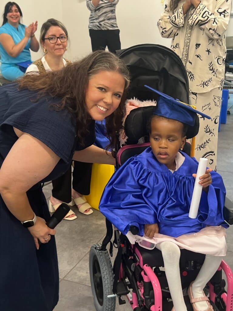 A woman smiles beside a young girl in a wheelchair wearing a blue graduation cap and gown, holding a rolled-up certificate. Several people in the background are watching and clapping.
