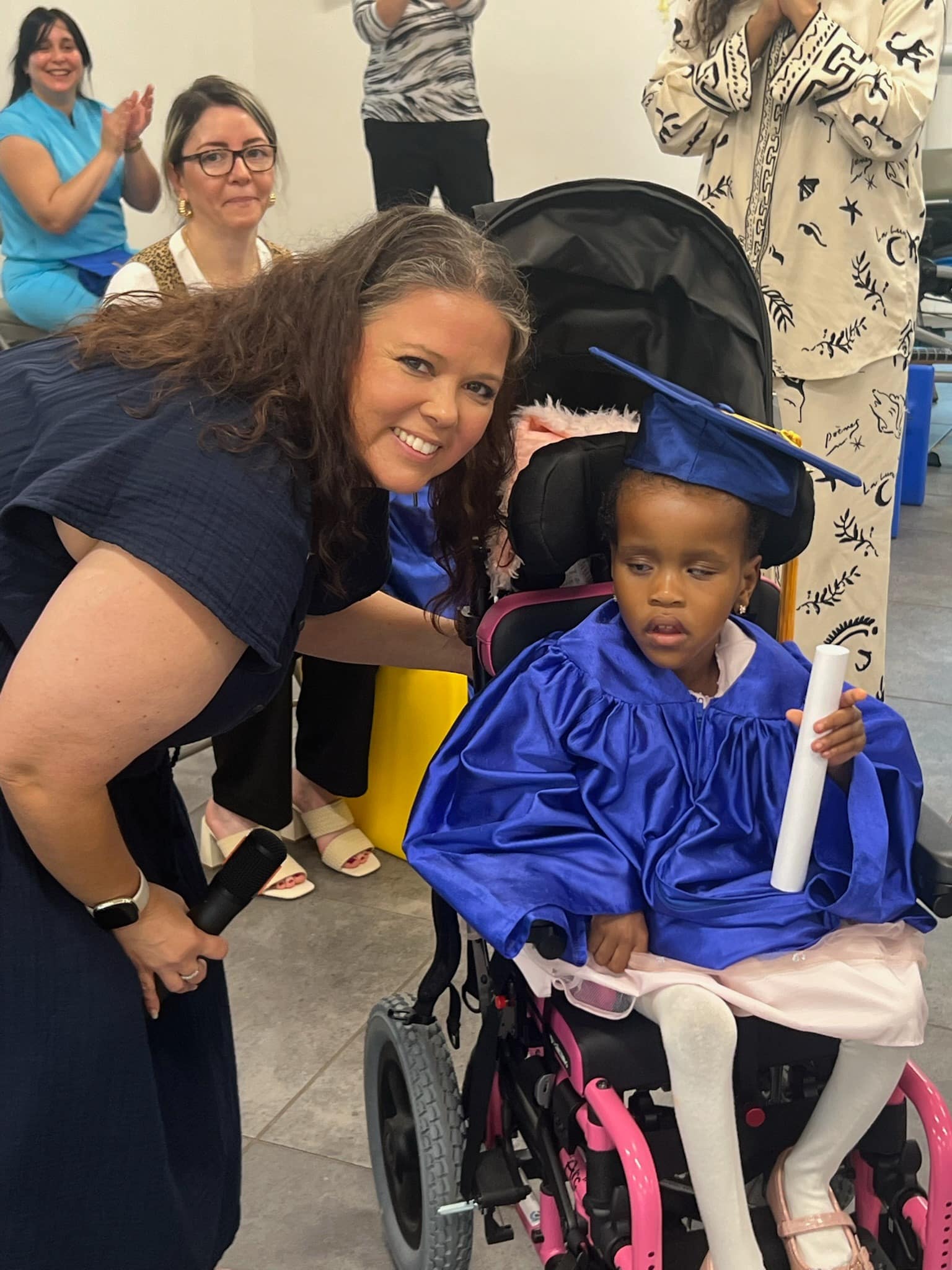 A woman smiles beside a young girl in a wheelchair wearing a blue graduation cap and gown, holding a rolled-up certificate. Several people in the background are watching and clapping.