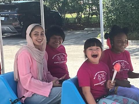 A woman in a light pink hijab and three smiling children in matching pink shirts sit together on a blue bench outdoors, enjoying a sunny day. One child holds a pink toy. Trees and parked cars are in the background.