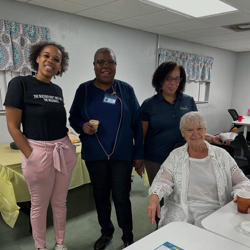 Four women pose together in a bright room. Three stand behind a seated woman dressed in white. A table with food and a yellow tablecloth is nearby. The atmosphere appears friendly and casual.