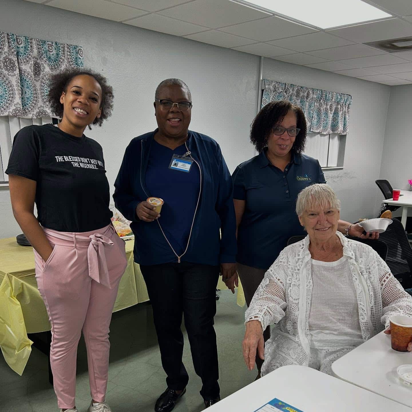 Four women pose together in a bright room. Three stand behind a seated woman dressed in white. A table with food and a yellow tablecloth is nearby. The atmosphere appears friendly and casual.