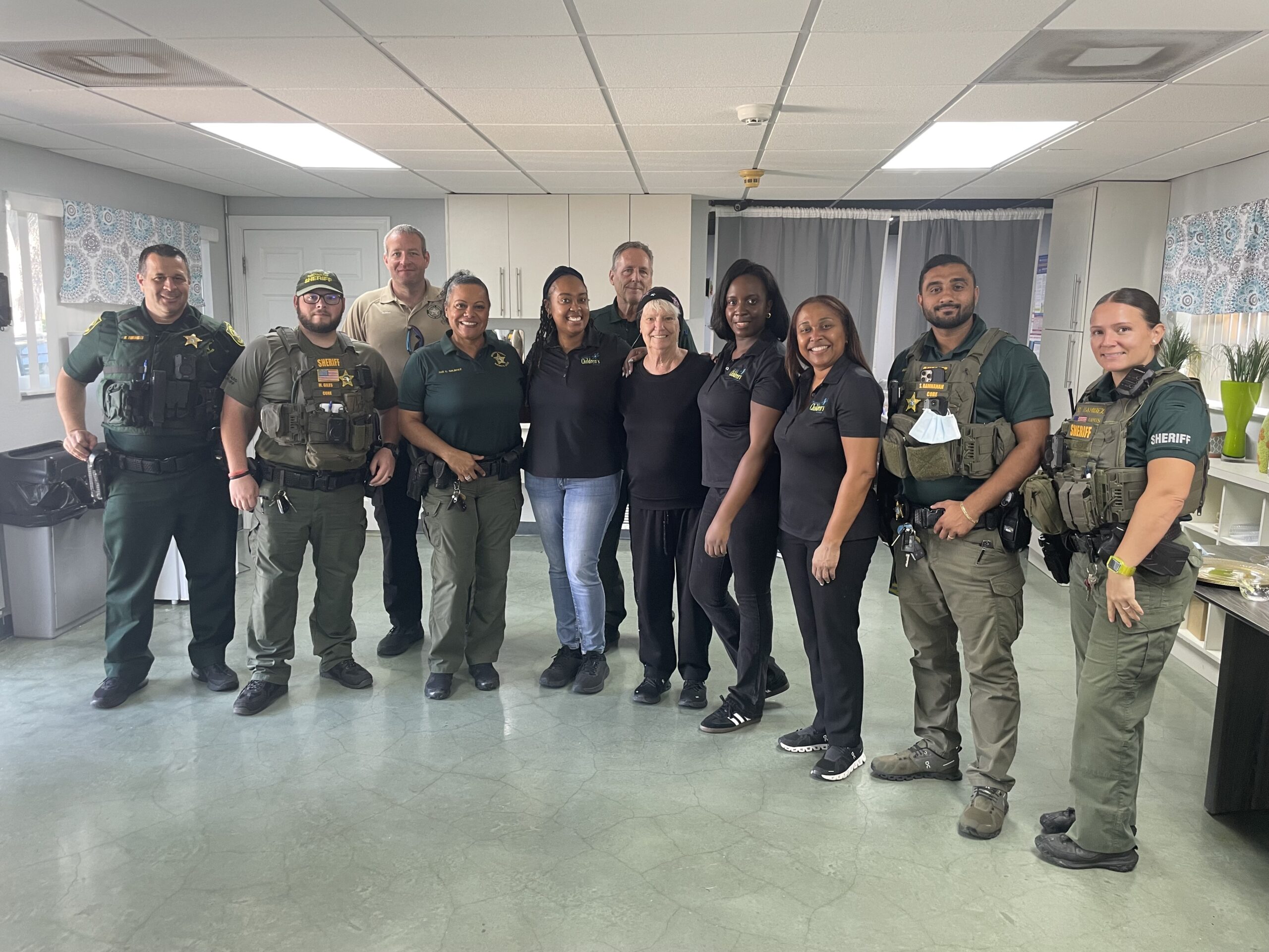 A group of twelve people, including security officers in uniform and others in black outfits, stand together smiling in a room with light-colored walls, windows, and kitchen appliances.