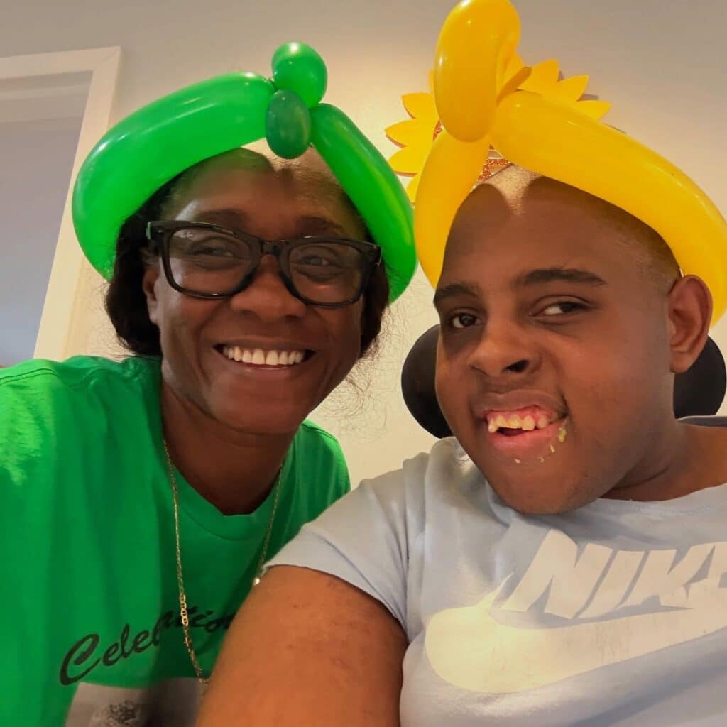 Two people smiling together indoors, both wearing colorful balloon hats—one green and one yellow. The person on the left wears glasses and a green shirt; the person on the right wears a light blue Nike shirt.