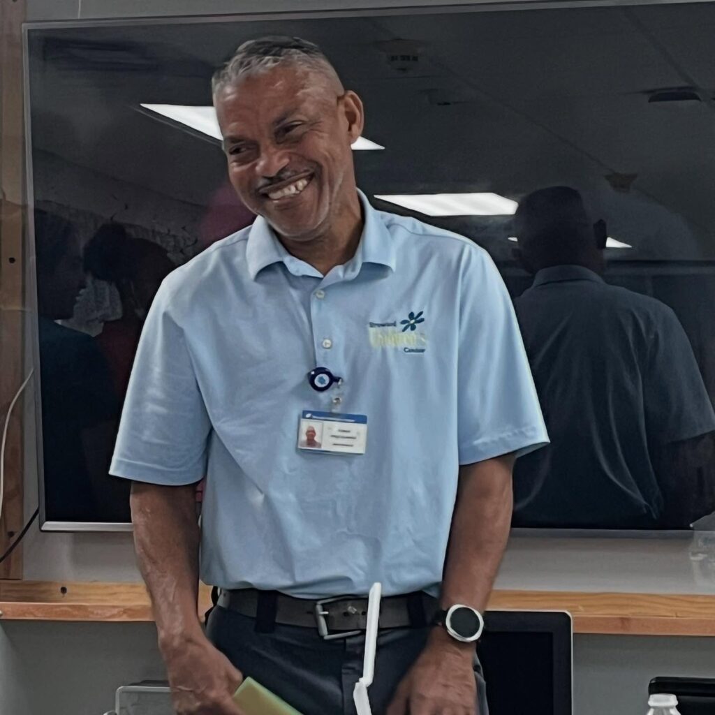 A smiling man wearing a light blue collared shirt with a company logo, a name badge, and a round watch stands in front of a large TV screen, appearing to interact in a professional or classroom setting.